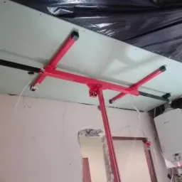 Czerwony podnośnik do płyt kartonowo-gipsowych podtrzymuje płytę przymocowaną do sufitu, widoczne profile metalowe, ściany w trakcie remontu, w tle piec grzewczy.