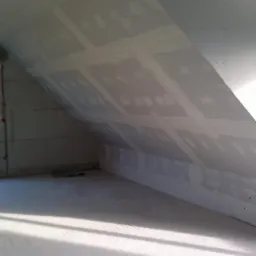 Poddasze w trakcie wykańczania, widoczne płyty kartonowo-gipsowe na skosach, częściowo zaszpachlowane łączenia, surowy stan podłogi, okno z widokiem na drzewa.