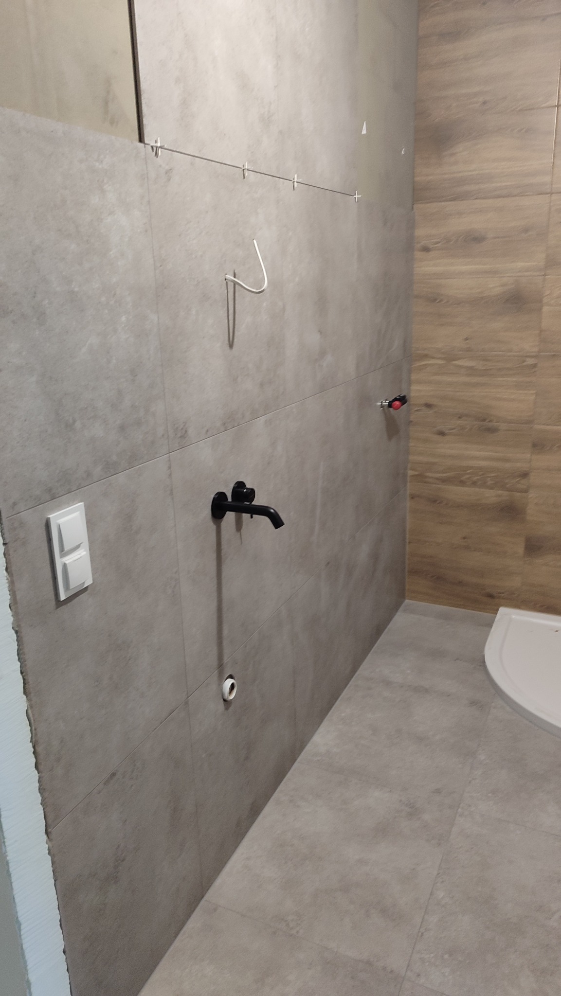 Wykończona ściana łazienki z szarych płytek, czarną armaturą i fragmentem toalety. Widoczne krzyżyki dystansowe i pionowy fragment drewnopodobnych płytek.