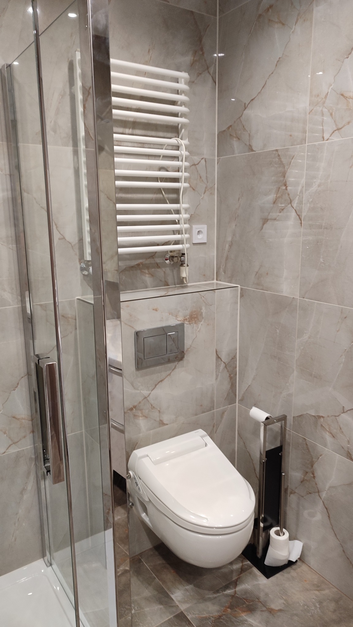 Elegancka łazienka po remoncie: marmurowe płytki, podwieszana toaleta z funkcją bidetu, chromowany grzejnik drabinkowy i szklana kabina prysznicowa.