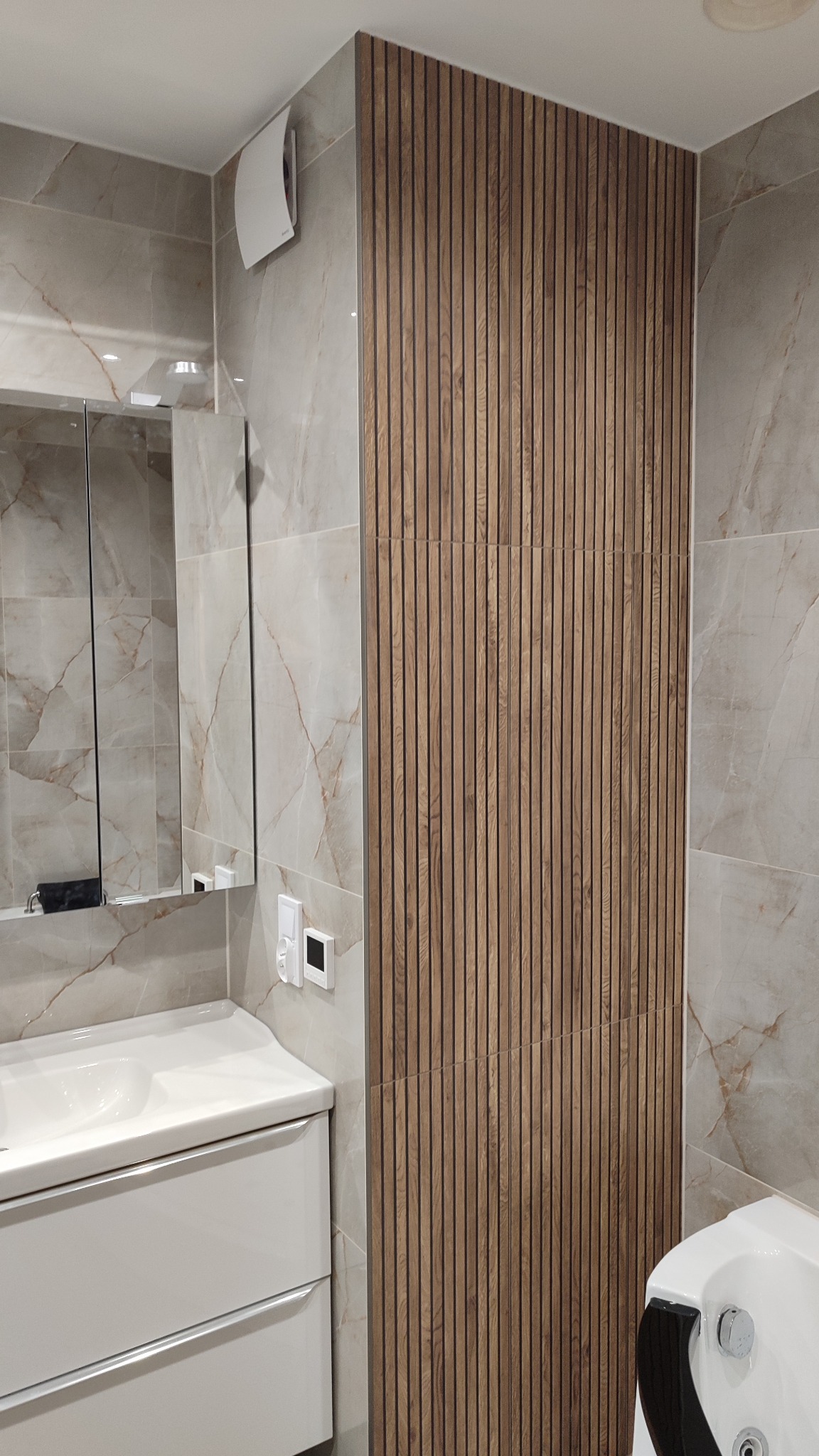 Wykończona łazienka z beżowymi płytkami imitującymi marmur i drewnianymi lamelami na ścianie. Widoczna biała umywalka z szafką i lustro.