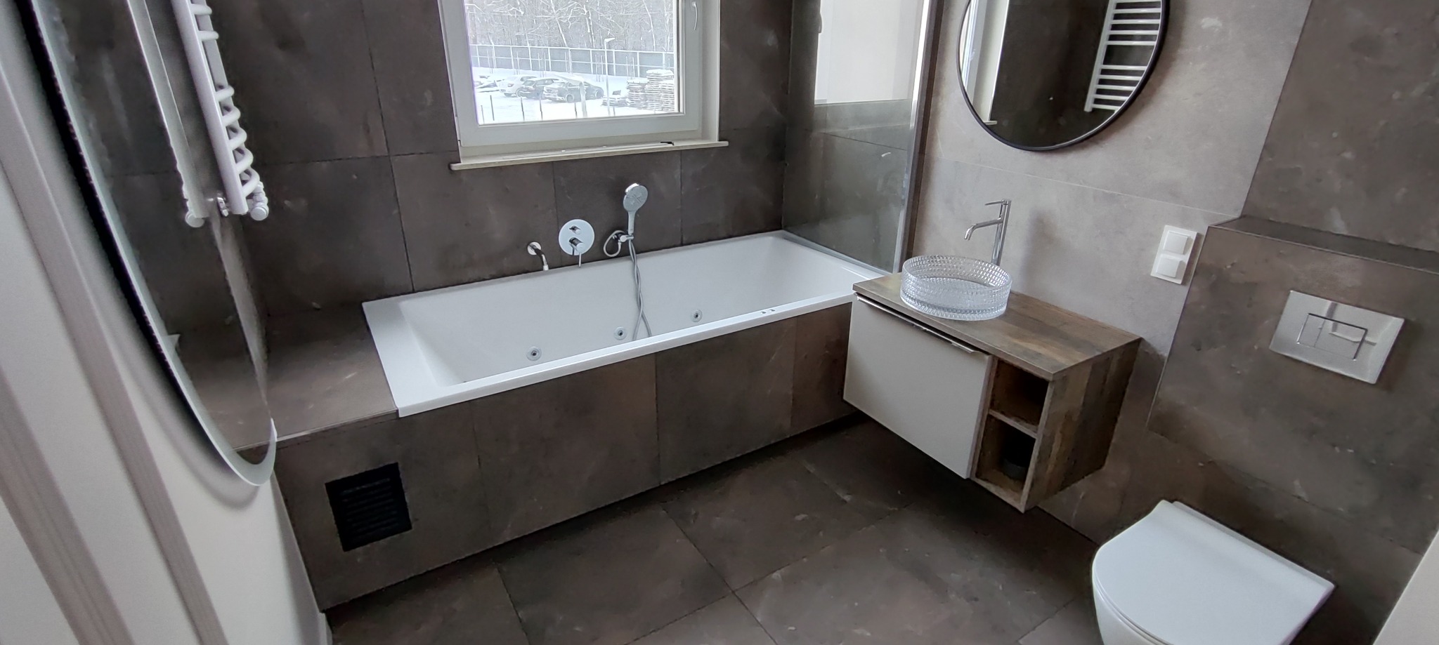 Nowoczesna łazienka z wanną jacuzzi, umywalką nablatową i szarą płytką. Widok na okno z zaśnieżonym krajobrazem za szybą. Minimalistyczny design z elementami drewna.