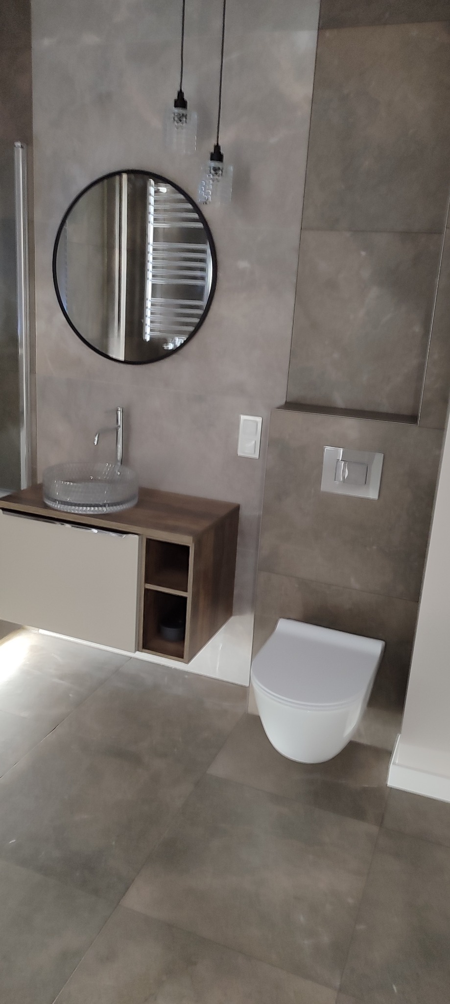 Nowoczesna łazienka w szarościach z okrągłym lustrem, szklaną umywalką i wiszącą toaletą. Minimalistyczne oświetlenie i płytki imitujące beton dodają elegancji.