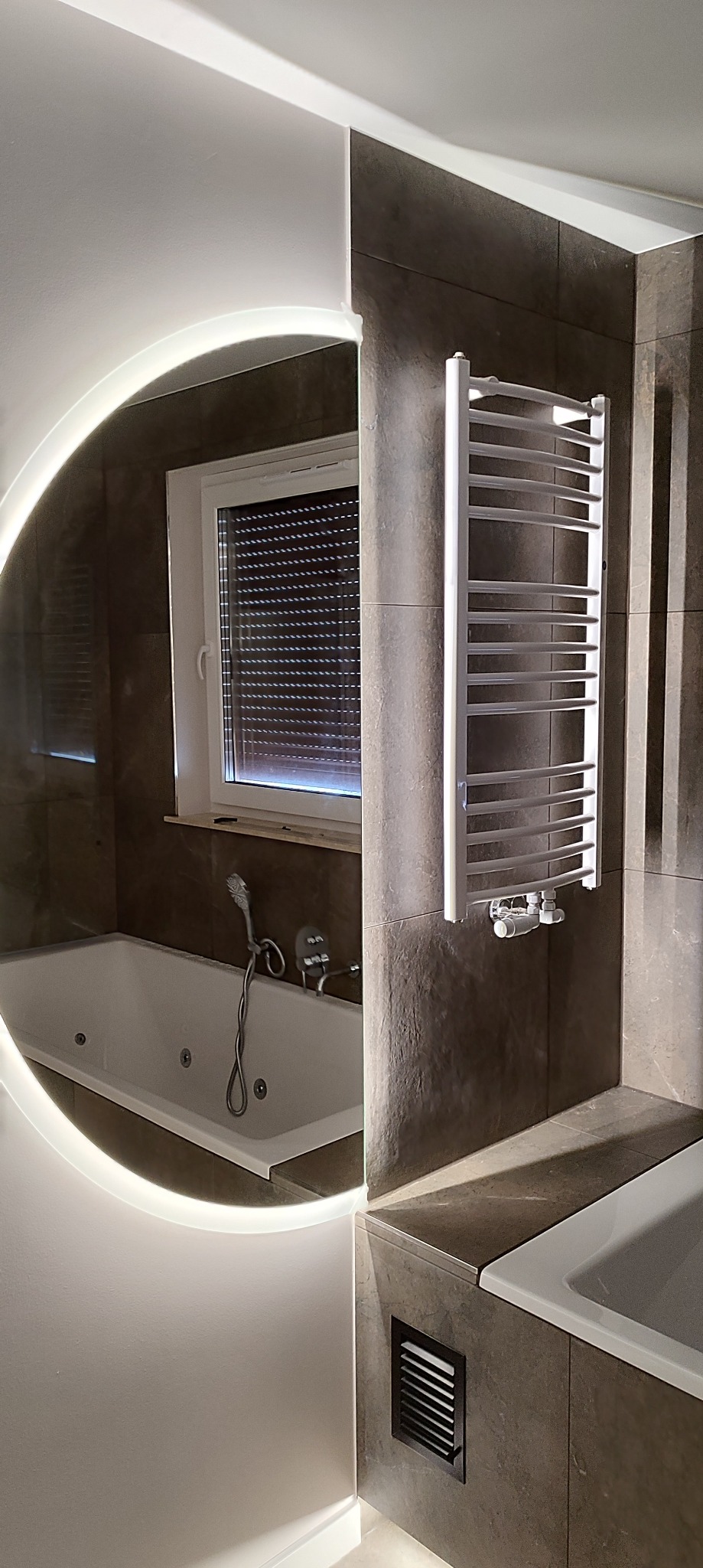 Wykończona łazienka z wanną z hydromasażem, grzejnikiem drabinkowym i okrągłym lustrem z podświetleniem LED. Ciemne płytki imitujące kamień.