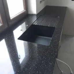 Czarny, błyszczący blat kuchenny z konglomeratu z wbudowanym, czarnym zlewozmywakiem jednokomorowym, widoczne otwory na baterię i dozownik, jasne odbicie światła w oknie.