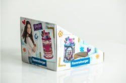 Ekspozytor reklamowy marki Ravensburger z grafiką przedstawiającą dziewczynę i motywy dekoracyjne, ustawiony na białym tle.