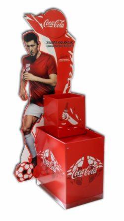 Stojak reklamowy z wizerunkiem Roberta Lewandowskiego i logo Coca-Cola, składający się z kartonowej figury piłkarza i dwóch czerwonych pudełek na produkty.