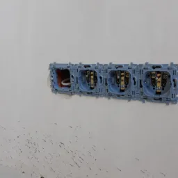 Szereg gniazdek elektrycznych podtynkowych w niebieskiej ramce, zamontowanych w surowej ścianie z widocznymi przewodami w pierwszym gniazdku po lewej stronie.