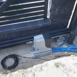 Szary automat do bramy przesuwnej zamontowany na betonowej podstawie, widoczne niebieskie rury osłonowe i zwinięty kabel zasilający na piaszczystym podłożu, brama z poziomych profili oraz fragment...