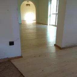 Jasna, surowa podłoga z desek w trakcie cyklinowania, widoczne ślady po szlifowaniu i fragment starej, brązowej podłogi w przejściu do innego pomieszczenia.