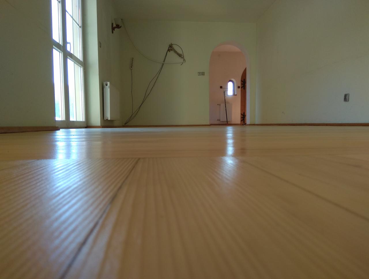 Jasna, lakierowana podłoga z desek w pustym pomieszczeniu, widoczny fragment okna z prawej strony i przejście do innego pokoju w tle.