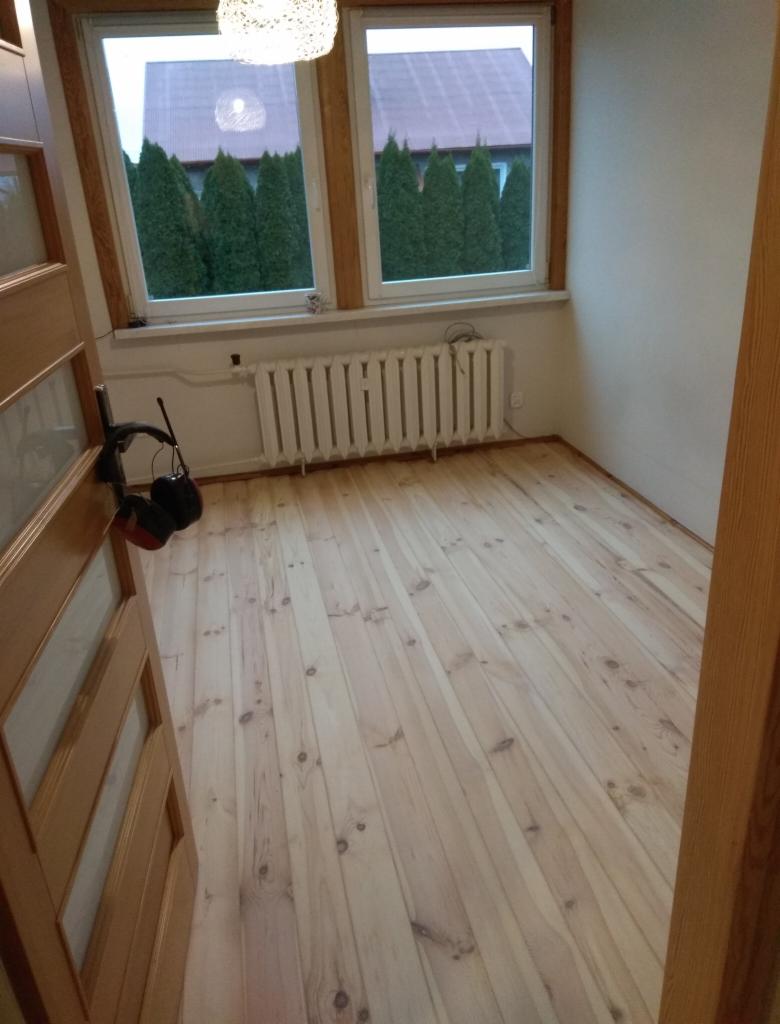 Jasna, surowa podłoga z desek w pokoju z oknem i grzejnikiem; słuchawki ochronne leżą przy drzwiach.