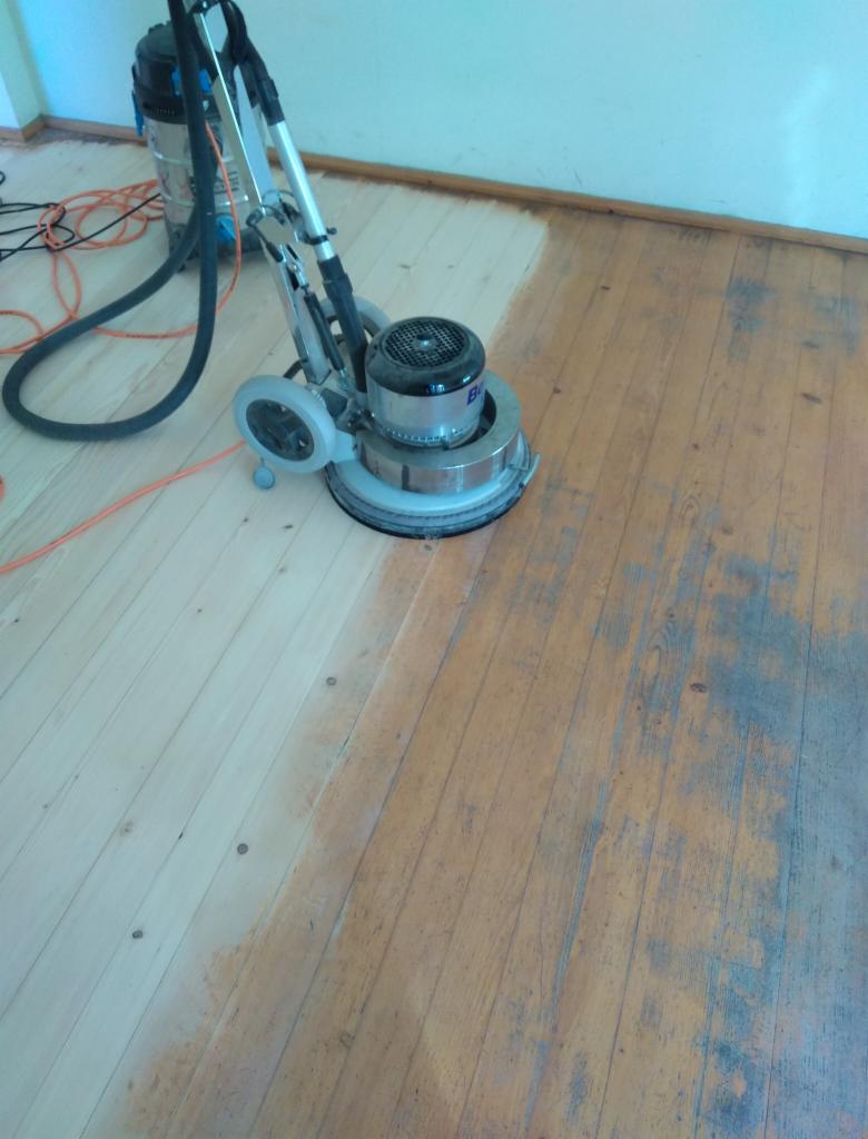Parkiet drewniany częściowo odnowiony przy użyciu szlifierki podłogowej, widoczna różnica w kolorze i fakturze między obszarem obrobionym a nieobrobionym, w tle odkurzacz przemysłowy.