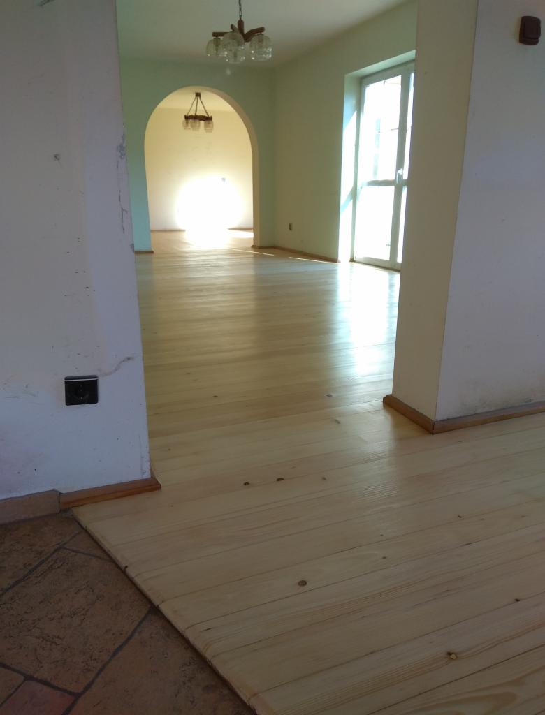 Jasna, surowa podłoga z desek w trakcie cyklinowania, widoczne ślady po szlifowaniu i fragment starej, brązowej podłogi w przejściu do innego pomieszczenia.