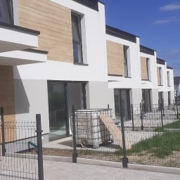 Nowoczesne szeregówki z elewacją w bieli i drewnie, ogrodzone metalową siatką. Widoczne detale wykończenia i otoczenie budynków w słoneczny dzień.