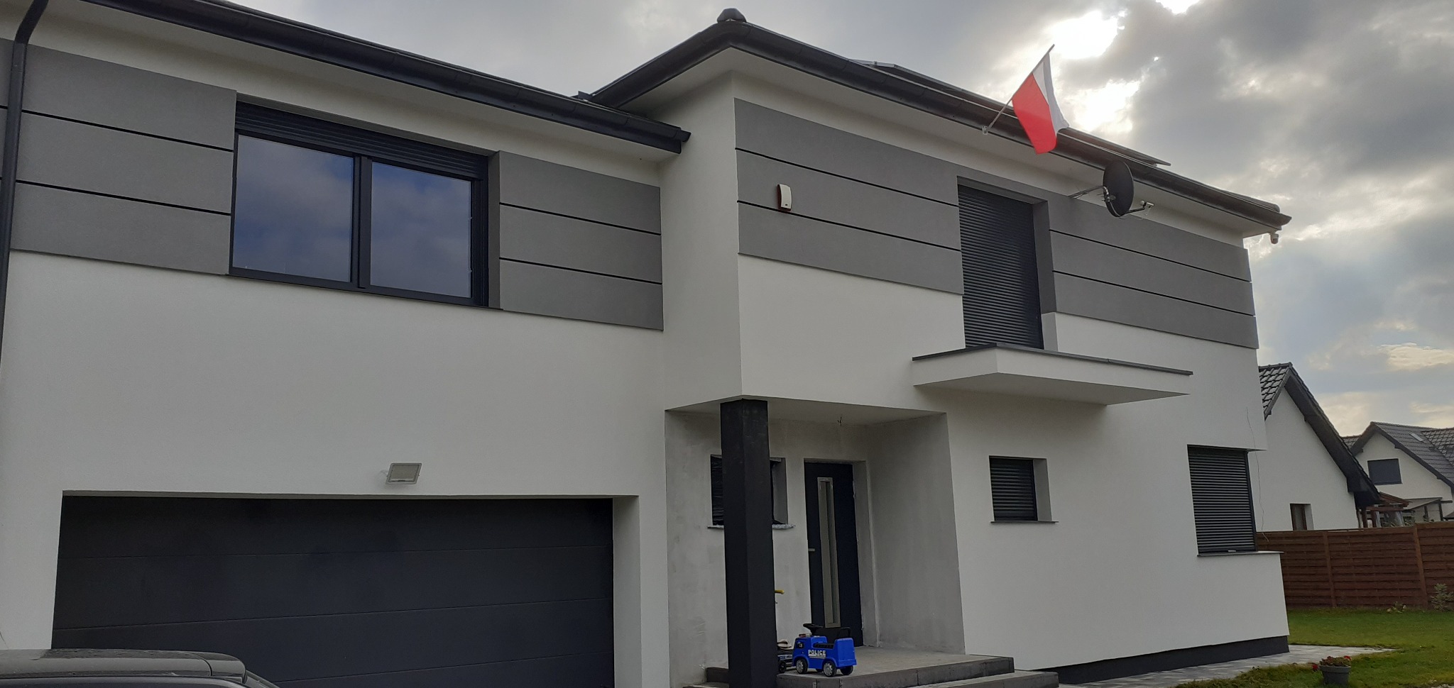 Nowoczesny dom z biało-szarą elewacją, czarnymi roletami i garażem. Na dachu flaga Polski. U dołu zabawka policyjna. Pochmurne niebo w tle.