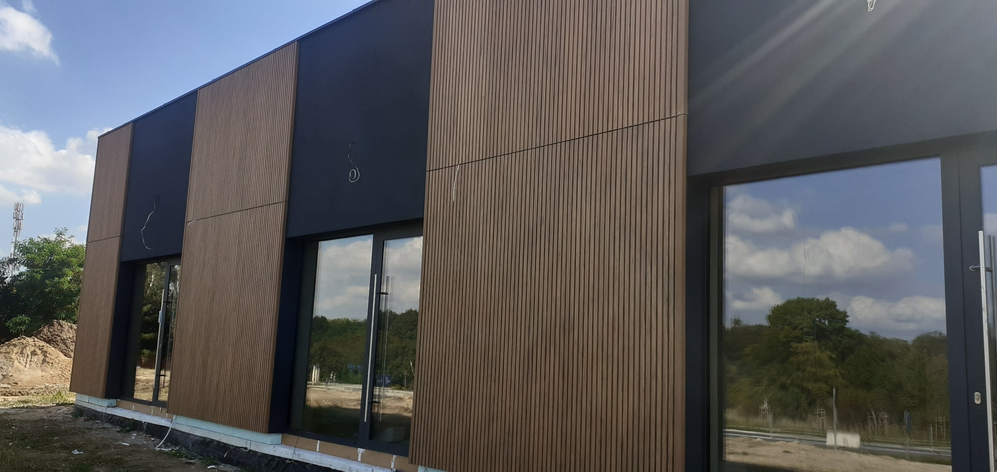 Nowoczesna elewacja budynku z brązowymi panelami i grafitowymi wstawkami. Widoczne okna z odbiciem zieleni. Częściowo wykończona podmurówka.