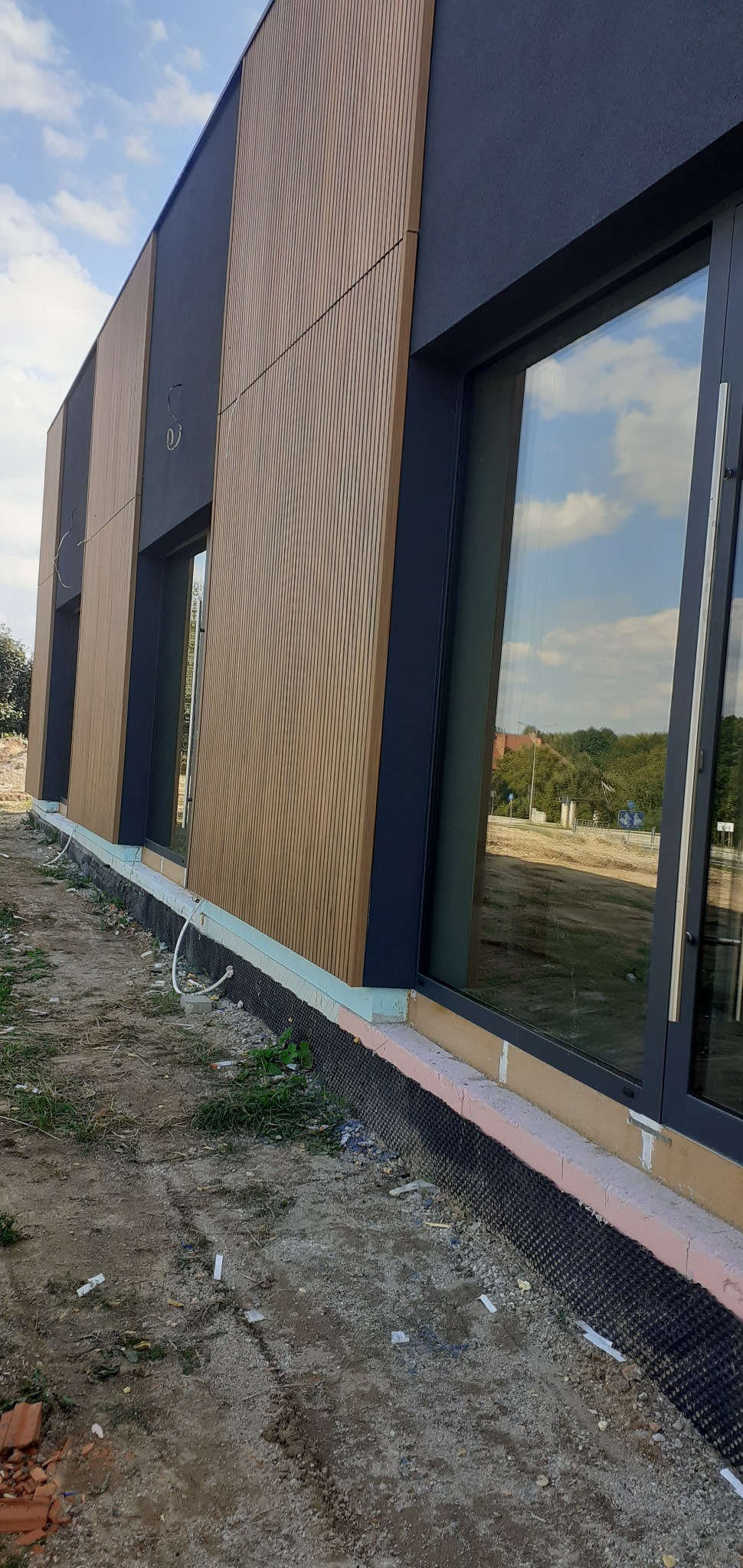 Nowoczesna elewacja budynku z drewnianymi panelami i grafitowymi wstawkami. Widoczne okna z odbiciem nieba i fragment terenu wokół budynku. Detal wykończenia.