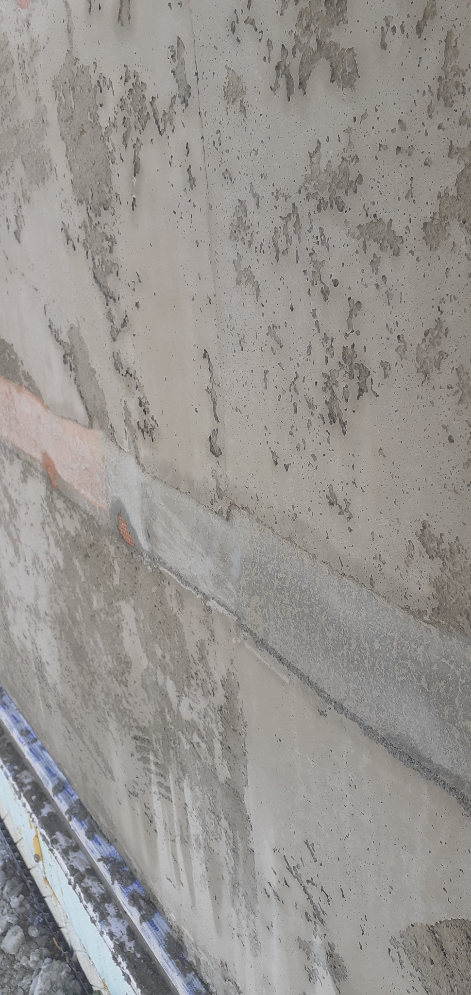 Szara, surowa ściana z widocznymi nierównościami i śladami po łataniu. W dolnej części kadru widoczne elementy rusztowania. Ujęcie z bliska, pod kątem.