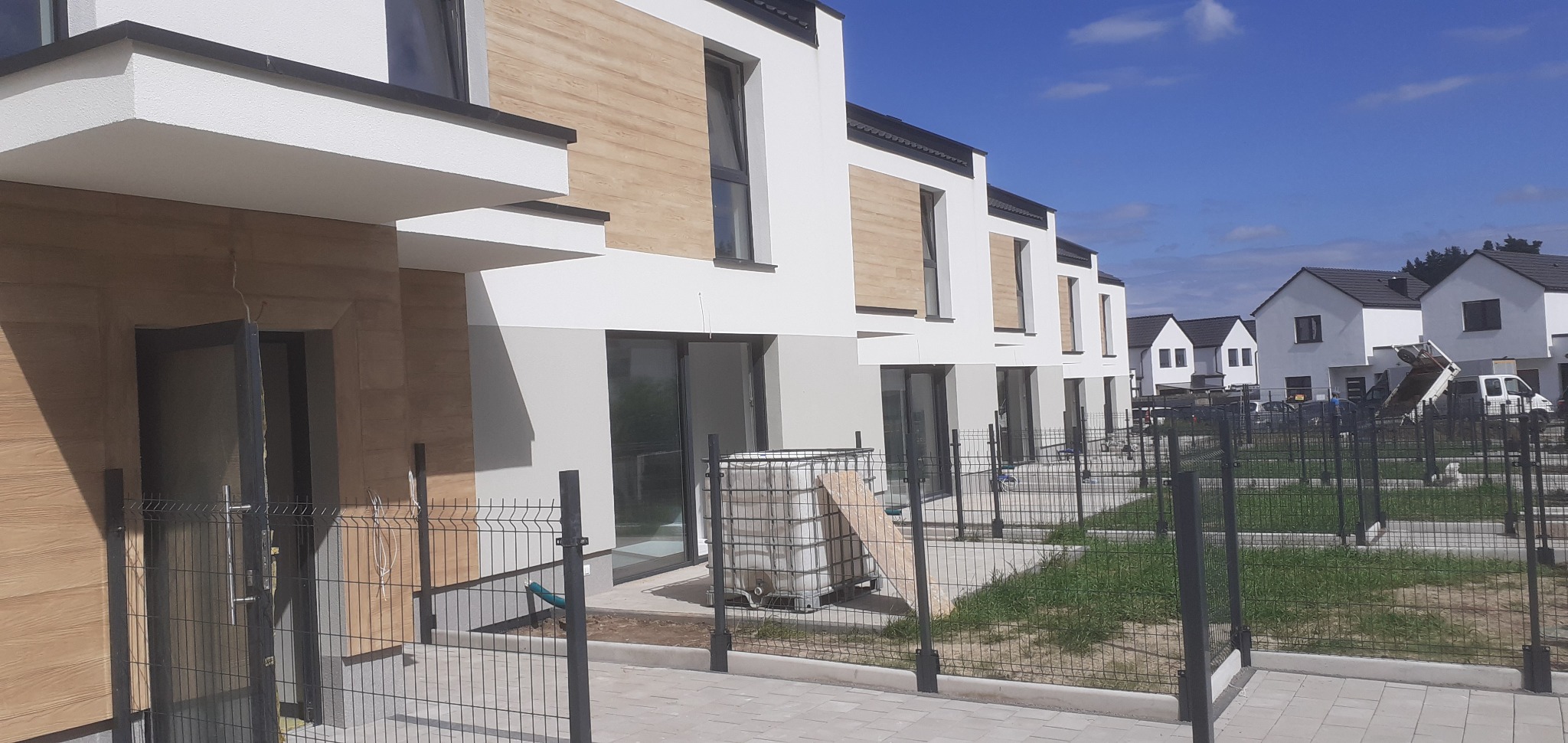 Nowoczesne szeregówki z elewacją w bieli i drewnie, ogrodzone metalową siatką. Widoczne detale wykończenia i otoczenie budynków w słoneczny dzień.