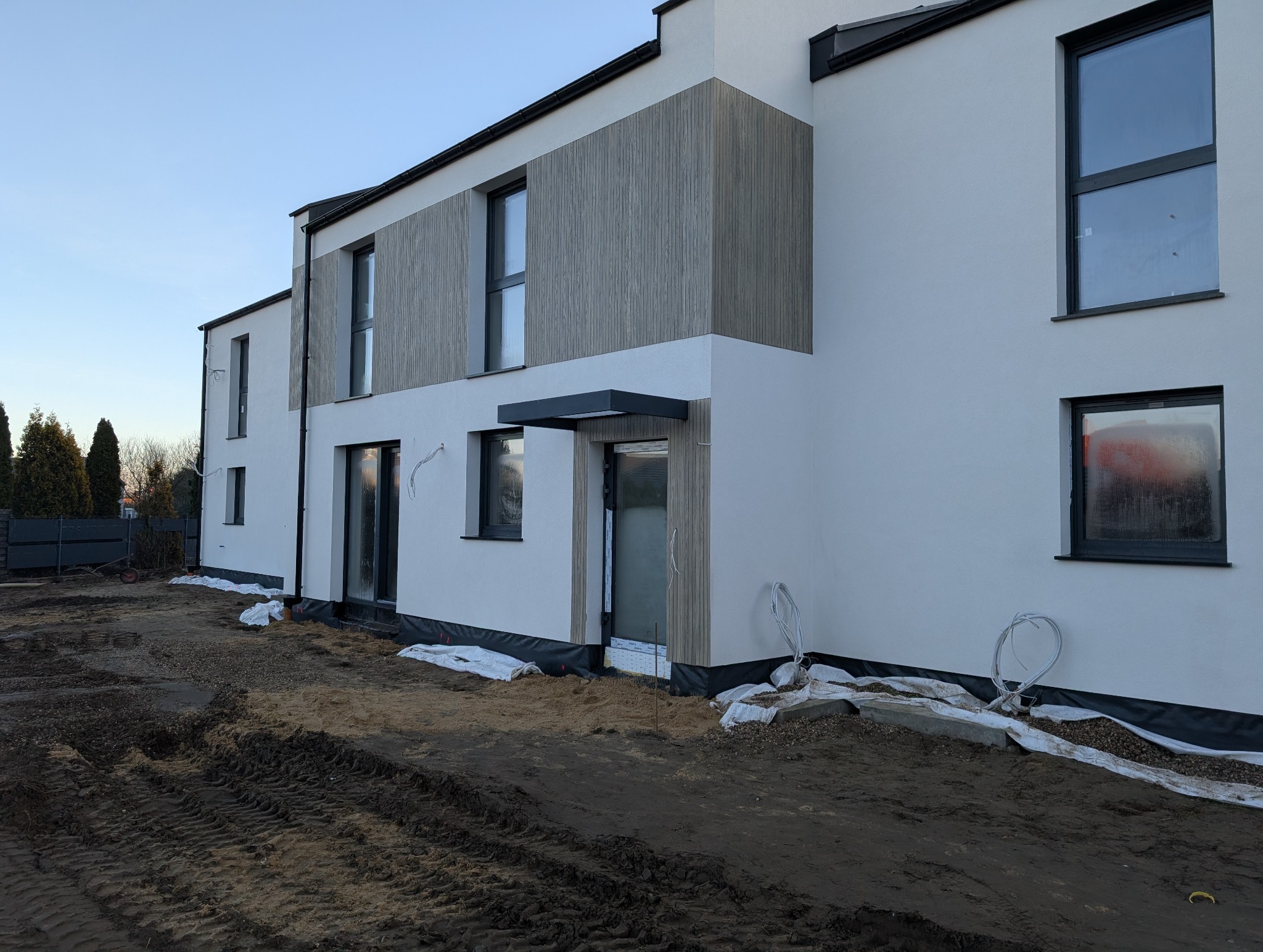 Nowoczesny dom z białą elewacją i drewnianymi panelami, widok z zewnątrz, teren wokół domu w trakcie prac wykończeniowych.