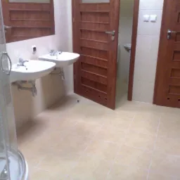 Wykończona łazienka z beżowymi płytkami, dwiema umywalkami i drewnianymi drzwiami z poziomymi panelami. Widoczna kabina prysznicowa i oświetlenie.