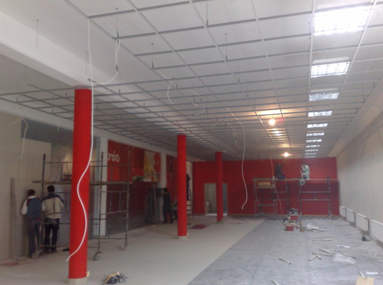 Montaż sufitu podwieszanego w lokalu komercyjnym z czerwonymi filarami i ścianami, widoczne rusztowania i pracownicy w trakcie pracy. Oświetlenie sufitowe częściowo zamontowane.