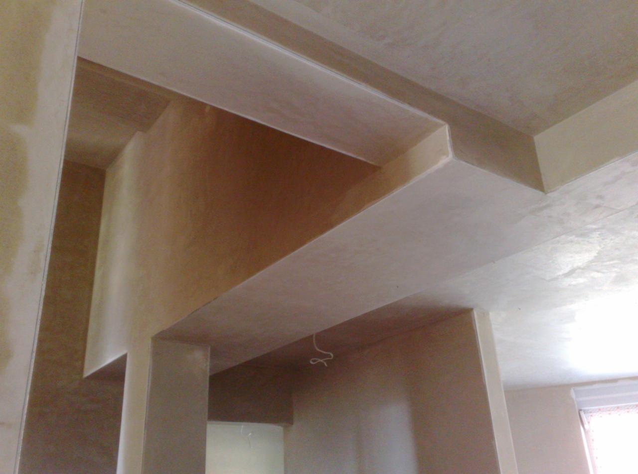 Świeżo położone tynki na ścianach i suficie z widocznymi detalami architektonicznymi wewnątrz pomieszczenia. Gładkie wykończenie, jasne kolory.