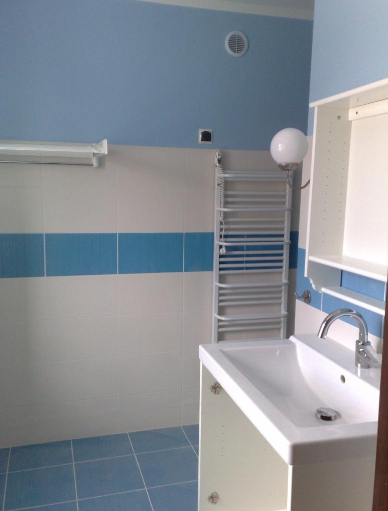 Wykończona łazienka w błękitno-białej tonacji z umywalką, grzejnikiem drabinkowym i płytkami. Widoczna wentylacja i oświetlenie. Ujęcie z niskiego kąta.