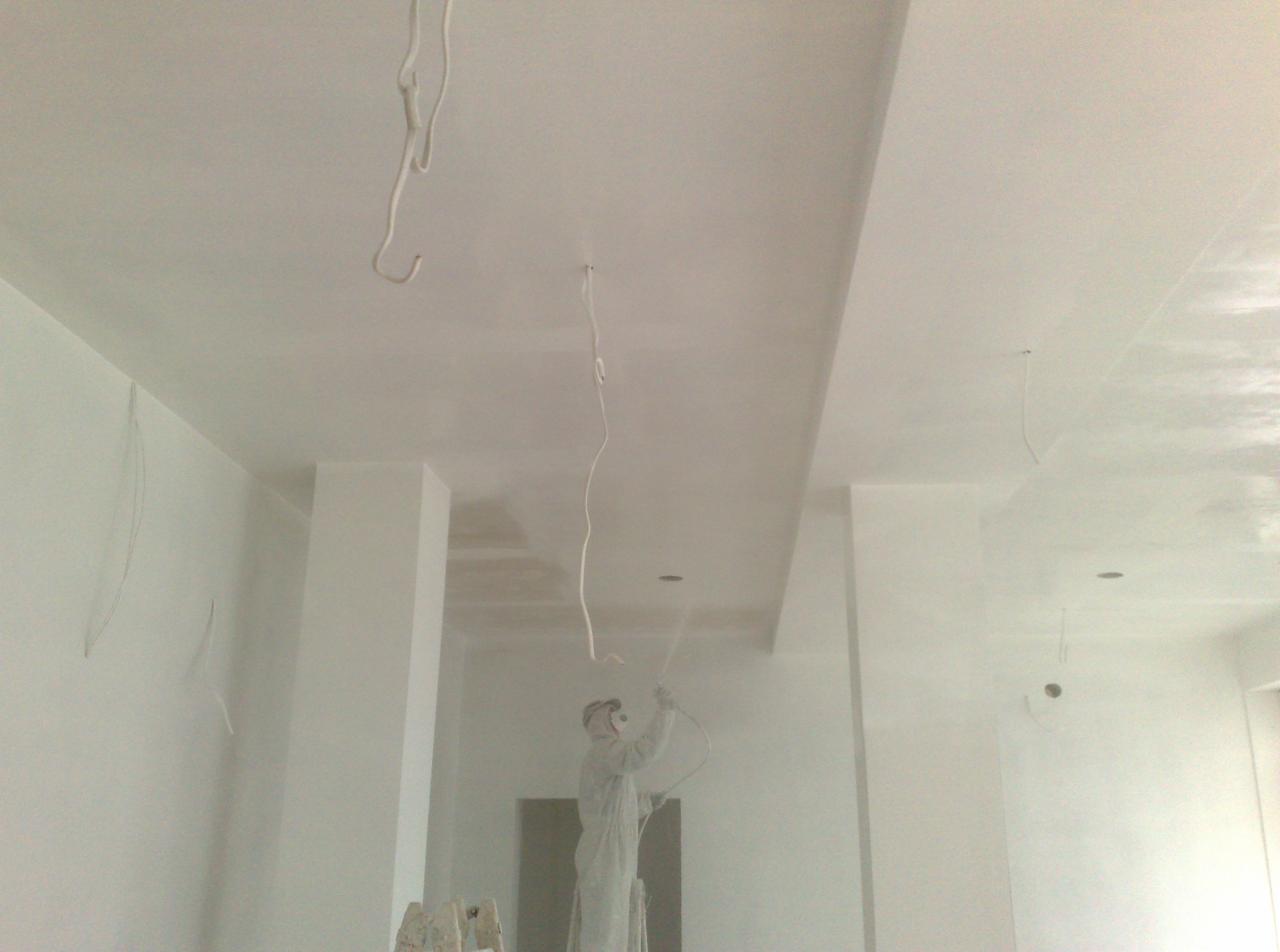 Pracownik w kombinezonie maluje natryskowo sufit w pomieszczeniu. Widoczne przewody zwisające z sufitu, kolumny i świeżo pomalowane ściany. Praca w toku.