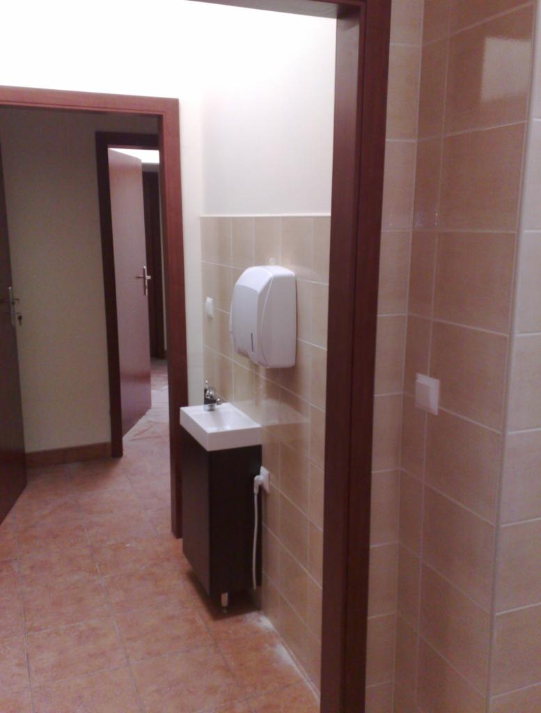 Beżowa łazienka z umywalką na szafce i dozownikiem mydła, widoczna przez otwarte drzwi z brązową ościeżnicą. Na podłodze i ścianach beżowe płytki.