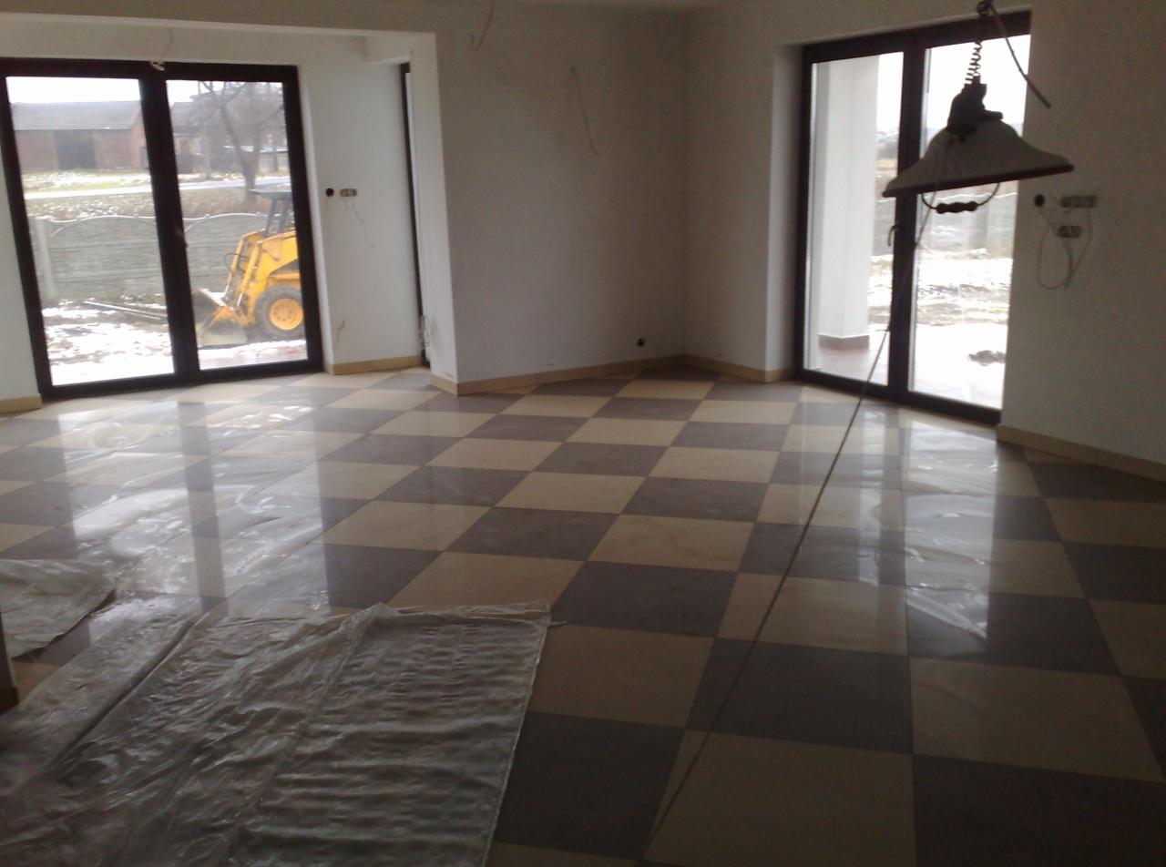 Szachownica z płytek podłogowych w jasnym wnętrzu z dużymi oknami. Część podłogi zabezpieczona folią ochronną. Na zewnątrz widoczny sprzęt budowlany.