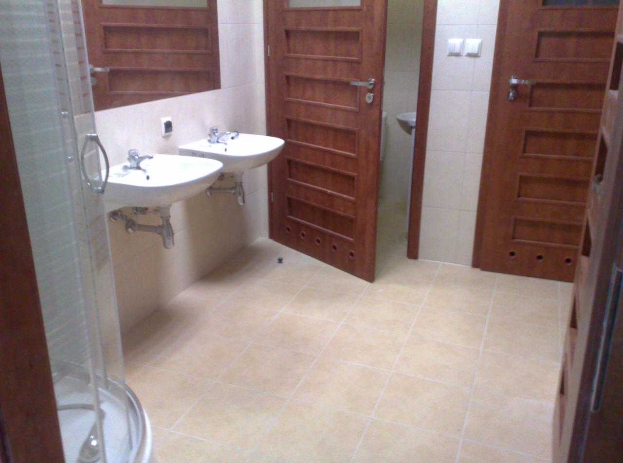 Wykończona łazienka z beżowymi płytkami, dwiema umywalkami i drewnianymi drzwiami z poziomymi panelami. Widoczna kabina prysznicowa i oświetlenie.