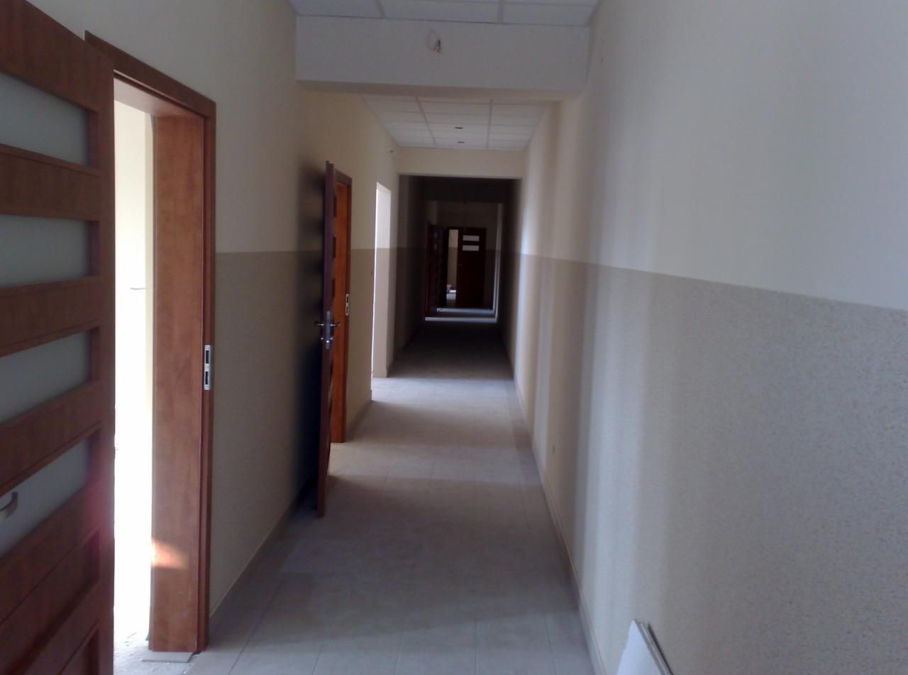 Długi korytarz z otwartymi drewnianymi drzwiami po lewej stronie, z dwukolorowymi ścianami w odcieniach beżu i szarości, prowadzący w głąb budynku.