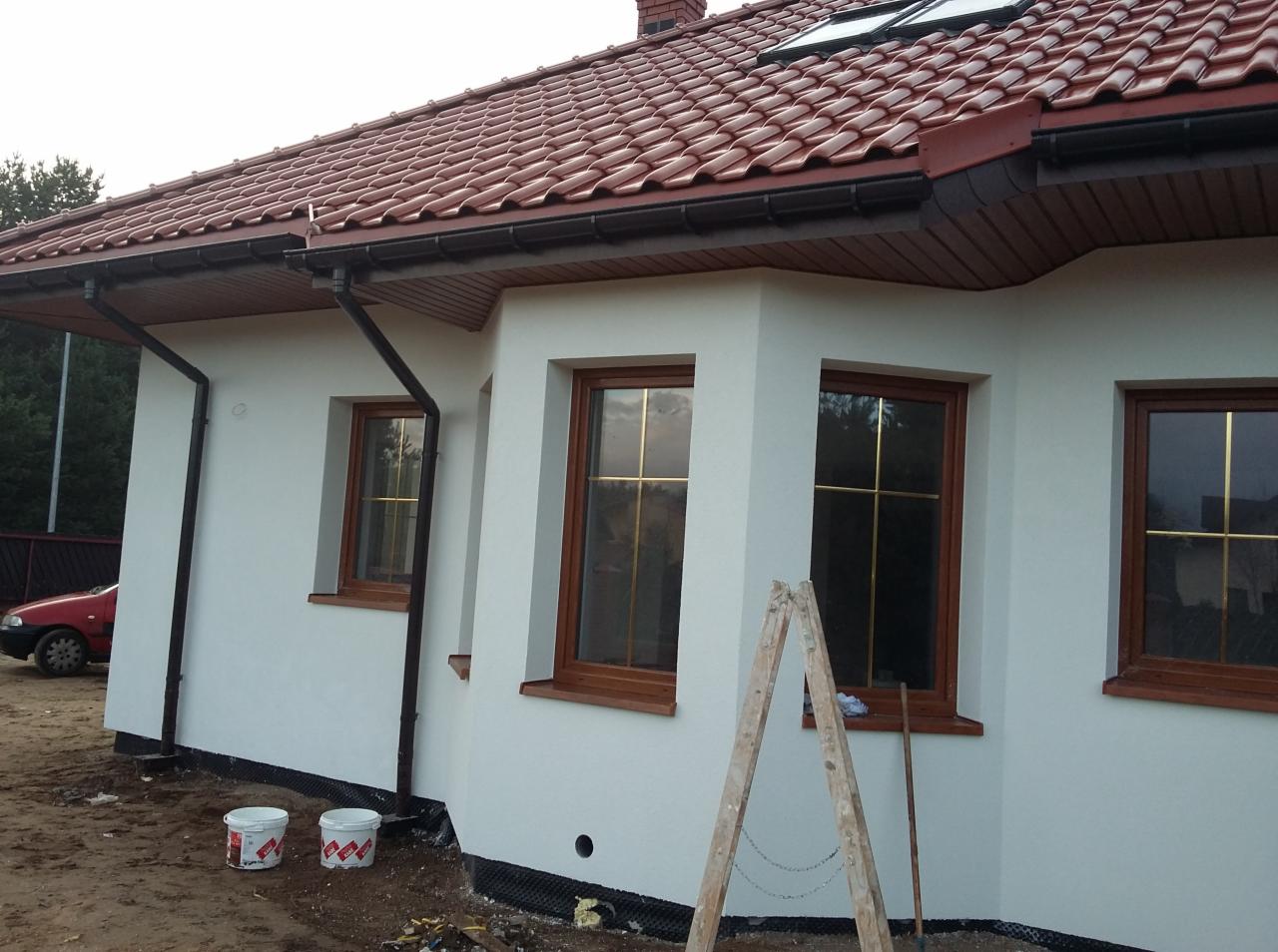 Świeżo otynkowany, biały dom z brązowymi oknami, widoczne dwie puszki z farbą i drabina. Czerwona dachówka i brązowe rynny uzupełniają wygląd zewnętrzny.