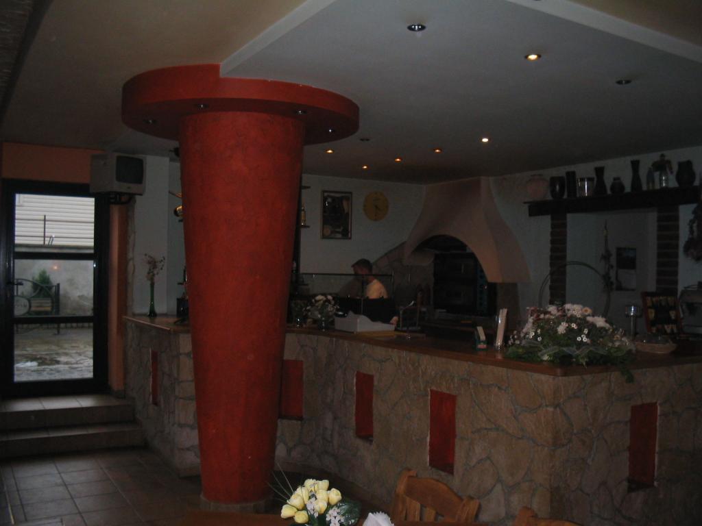 Wykończenie wnętrza lokalu z kolumną w kolorze czerwieni, kamienną ladą i widocznym piecem do pizzy w tle. Oświetlenie punktowe sufitowe.