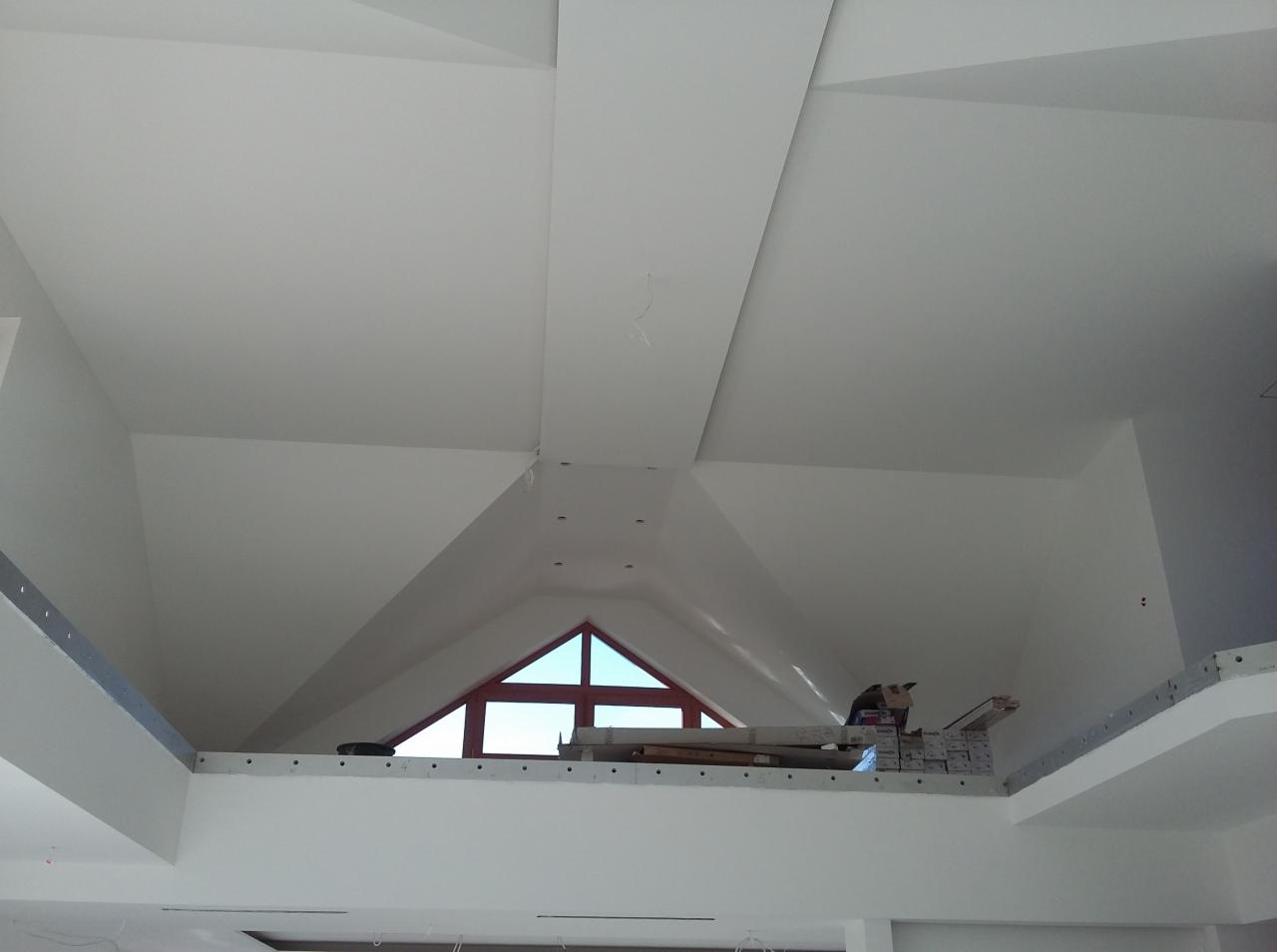 Białe, gładkie sufity pod skosem i ściany po remoncie, z widocznym trójkątnym oknem. Elementy konstrukcyjne i materiały budowlane na podeście.