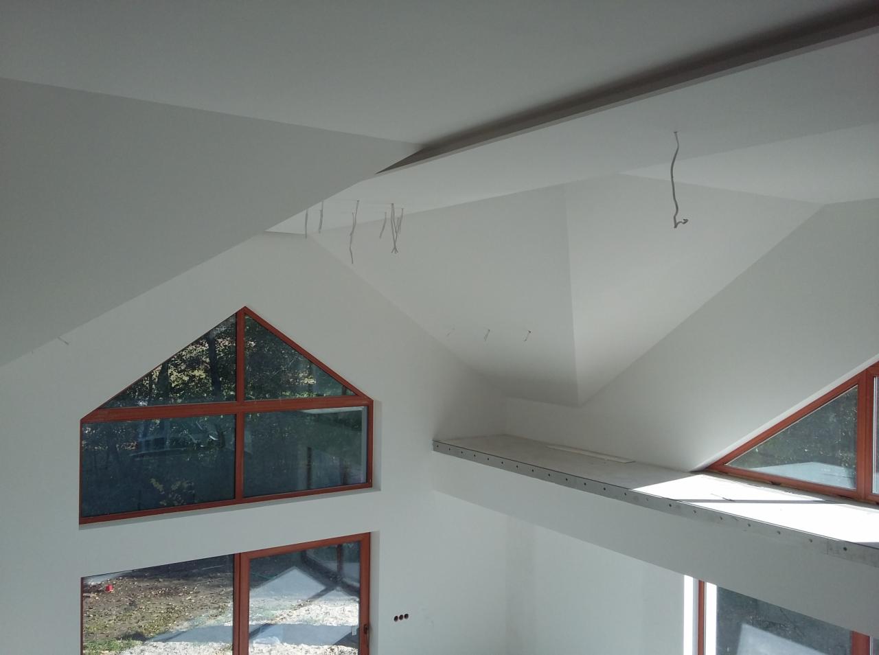 Wnętrze poddasza po tynkowaniu, z oknami o drewnianych ramach. Widoczne przewody wystające z sufitu. Świeżo położone, białe tynki na skośnych ścianach.