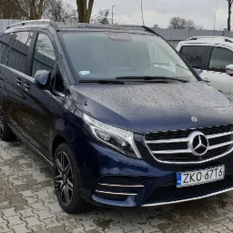 Granatowy Mercedes-Benz V-klasy z relingami dachowymi, zaparkowany na brukowanym placu w deszczowy dzień, widoczne tablice rejestracyjne ZKO 67T6.