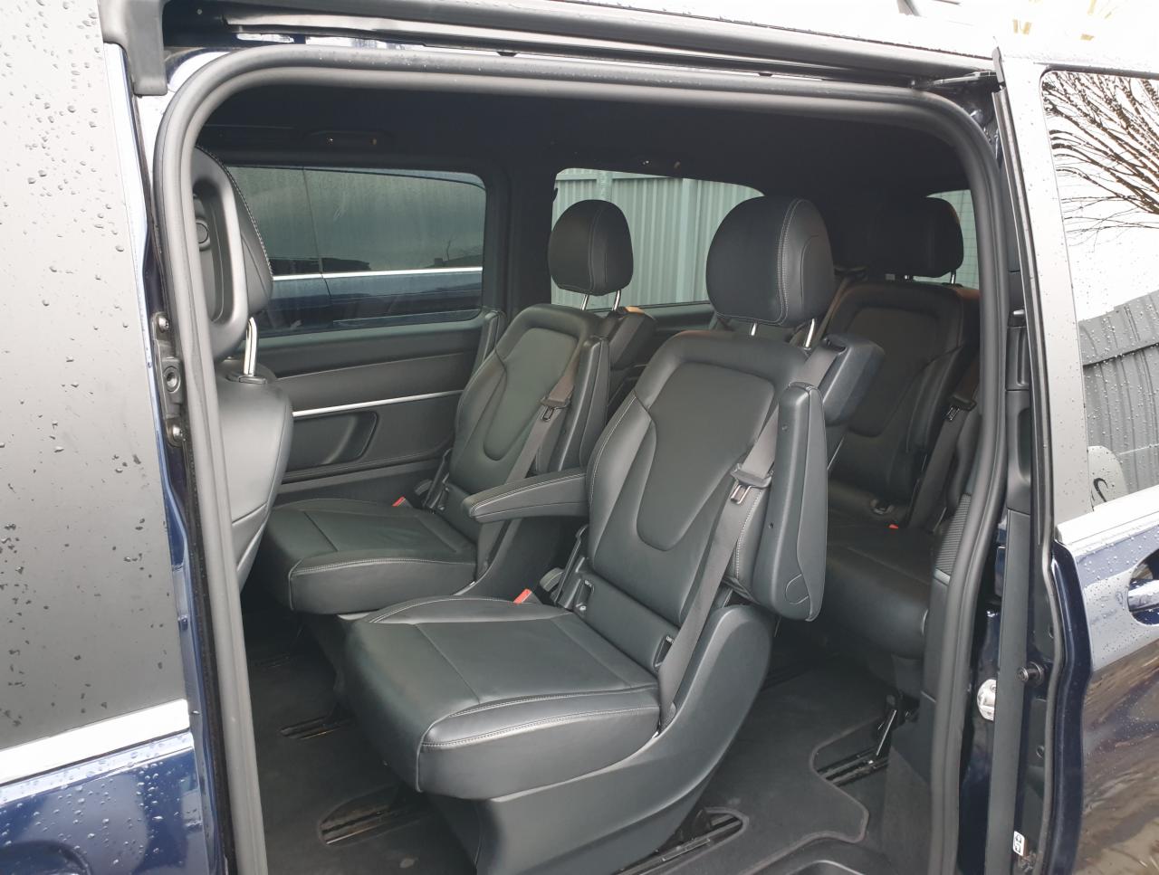 Wnętrze luksusowego busa z czarnymi skórzanymi fotelami, widok z otwartych drzwi bocznych, deszcz na szybach.