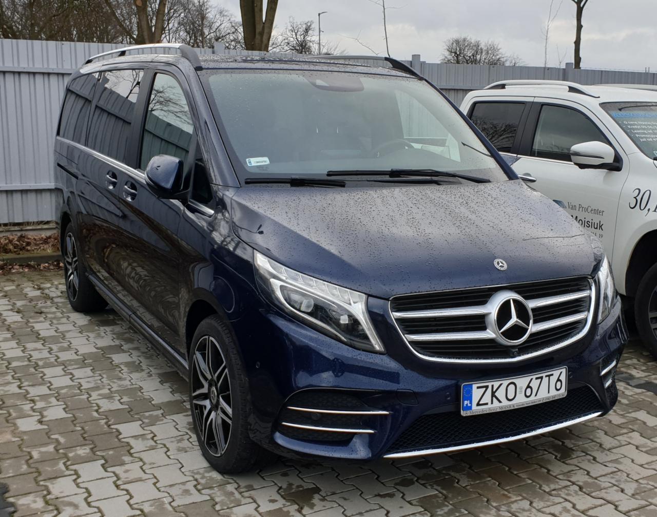 Granatowy Mercedes-Benz V-klasy z relingami dachowymi, zaparkowany na brukowanym placu w deszczowy dzień, widoczne tablice rejestracyjne ZKO 67T6.