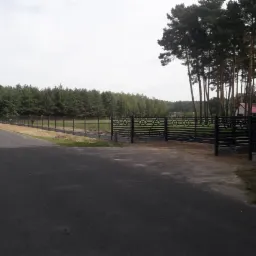 Nowoczesne ogrodzenie panelowe z bramą przesuwną i podmur&oacute;wką betonową, widoczne z perspektywy ulicy, w tle las i domy.