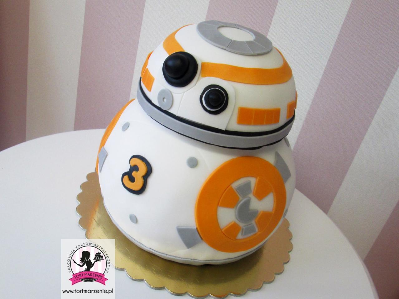 Tort w kształcie robota BB-8 z Gwiezdnych Wojen, z numerem 3, na złotym podkładzie, na białym stole, tło w różowe paski.