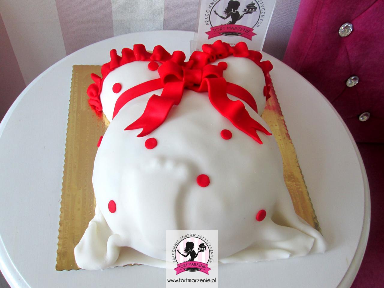 Tort w kształcie sukienki ciążowej z czerwonymi wstążkami i kropkami, wykonany w Tychach, na białym stole z logo firmy w tle.