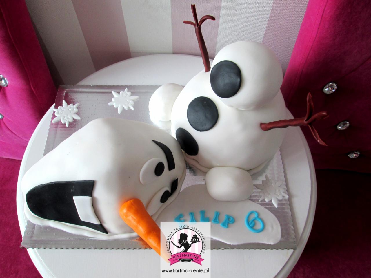 Tort w kształcie roztopionego Olafa z filmu 'Kraina Lodu', leżący na białym talerzu, z napisem 'Filip 6' wykonanym z niebieskiego lukru, na tle różowej kanapy z kryształkami.