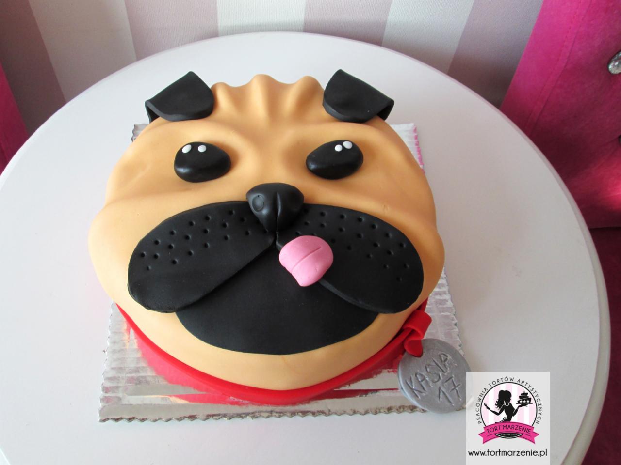 Tort w kształcie głowy mopsa z modeliny, z czarnym pyszczkiem, różowym językiem i srebrnym medalionem z napisem 'Kasia 17', na białym stole z różowymi paskami w tle.