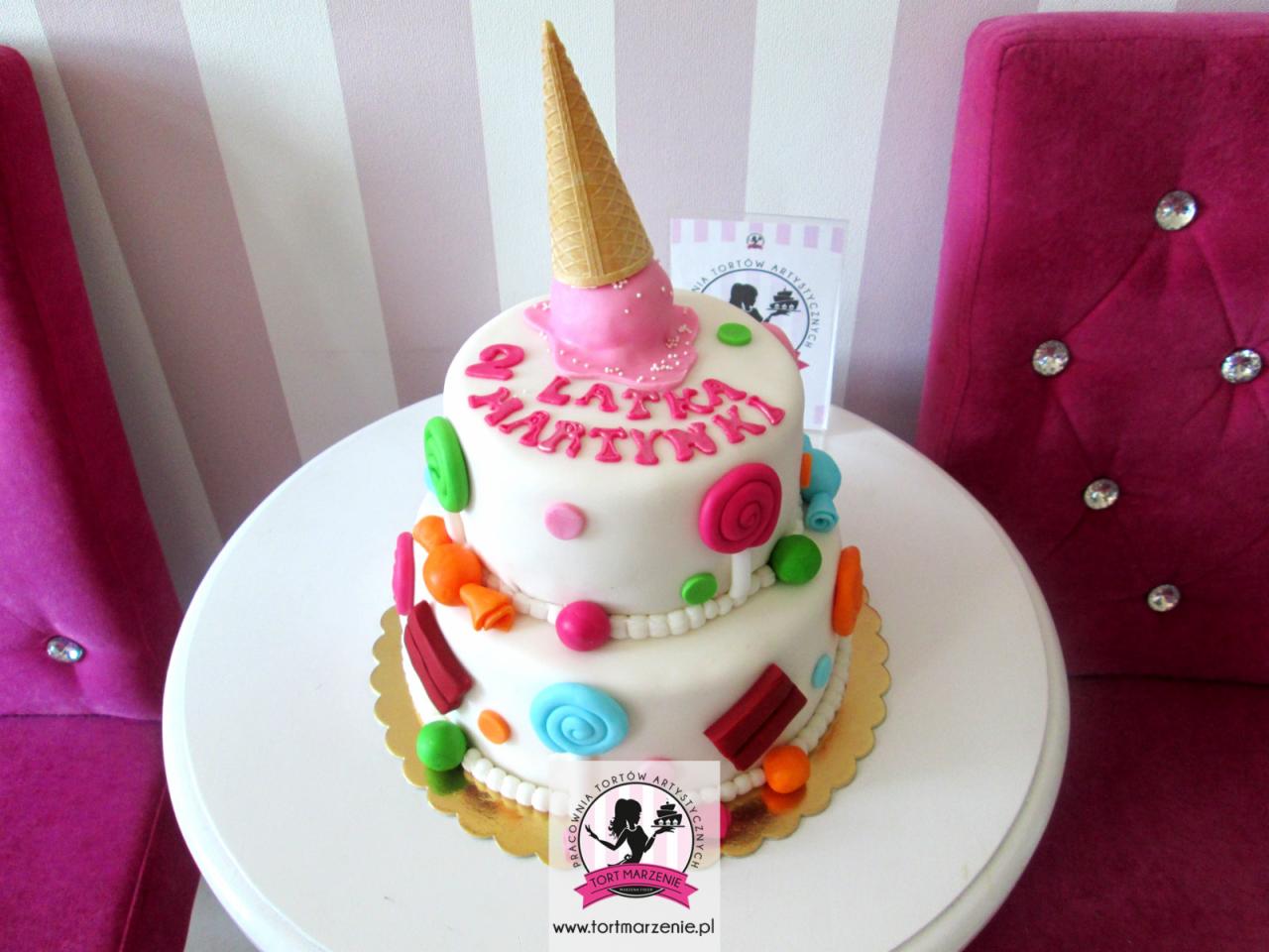 Dwupiętrowy tort urodzinowy z dekoracjami z lukru w kształcie cukierków i wafelkiem rożka na górze, dedykowany '2 Latka Martynki', na białym stoliku, z różowym krzesłem w tle.