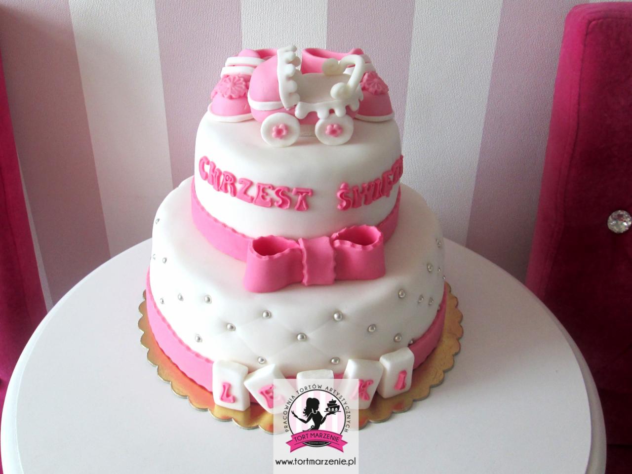 Dwupiętrowy tort na Chrzest Święty z różowymi dekoracjami: buciki, wózeczek, wstążka z kokardą, srebrne perełki i kostki z literami imienia dziecka.