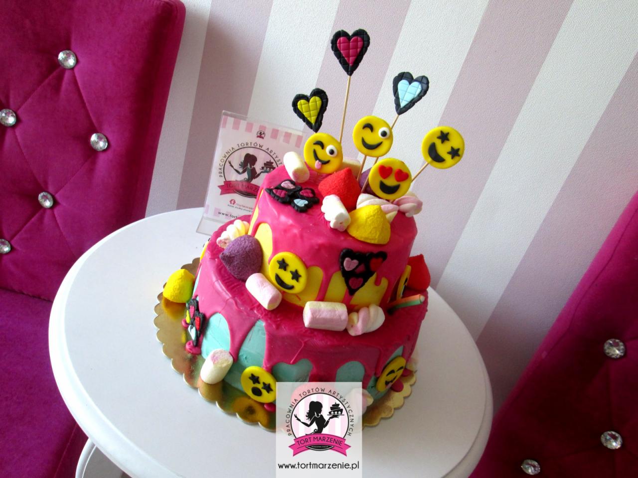 Dwupiętrowy tort w stylu emoji, z różowym lukrem spływającym po bokach, udekorowany piankami marshmallow, lizakami w kształcie emotikon i serduszek na patykach, stojący na złotym podkładzie, z logo...