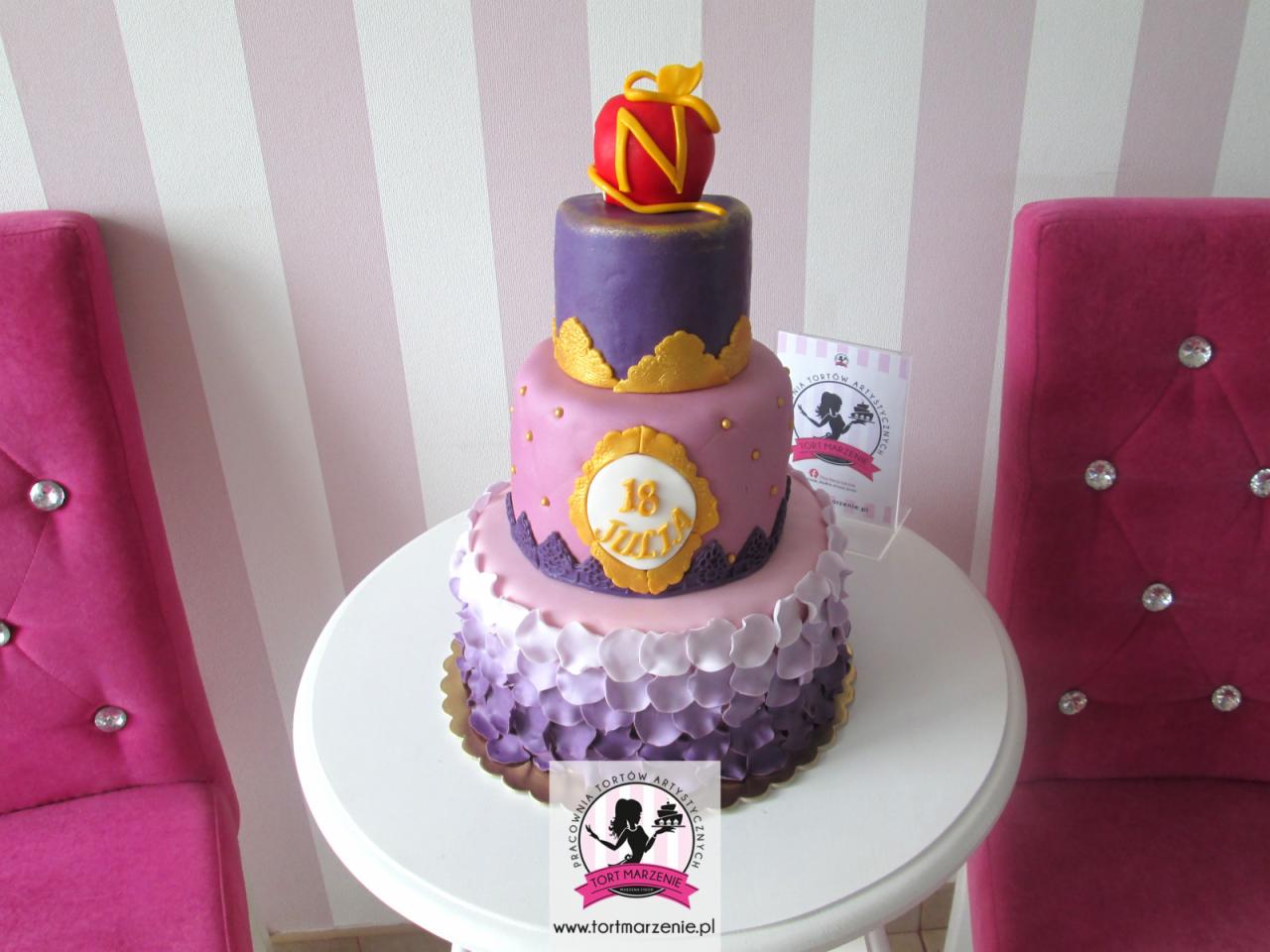 Wielopoziomowy tort urodzinowy w odcieniach fioletu i różu, zdobiony złotymi akcentami i czerwonym jabłkiem na szczycie, ustawiony na białym stoliku obok różowych krzeseł.